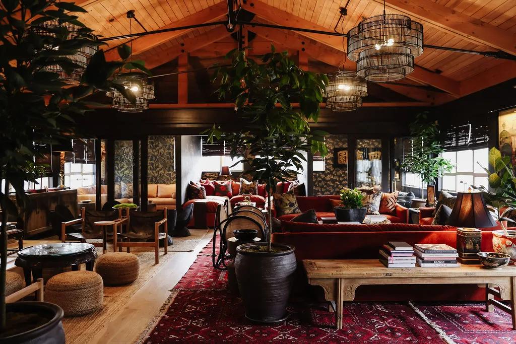 Clubhouse lounge viewed past potted trees with cage pendants, red rugs, velvet sofas and vaulted wood ceiling