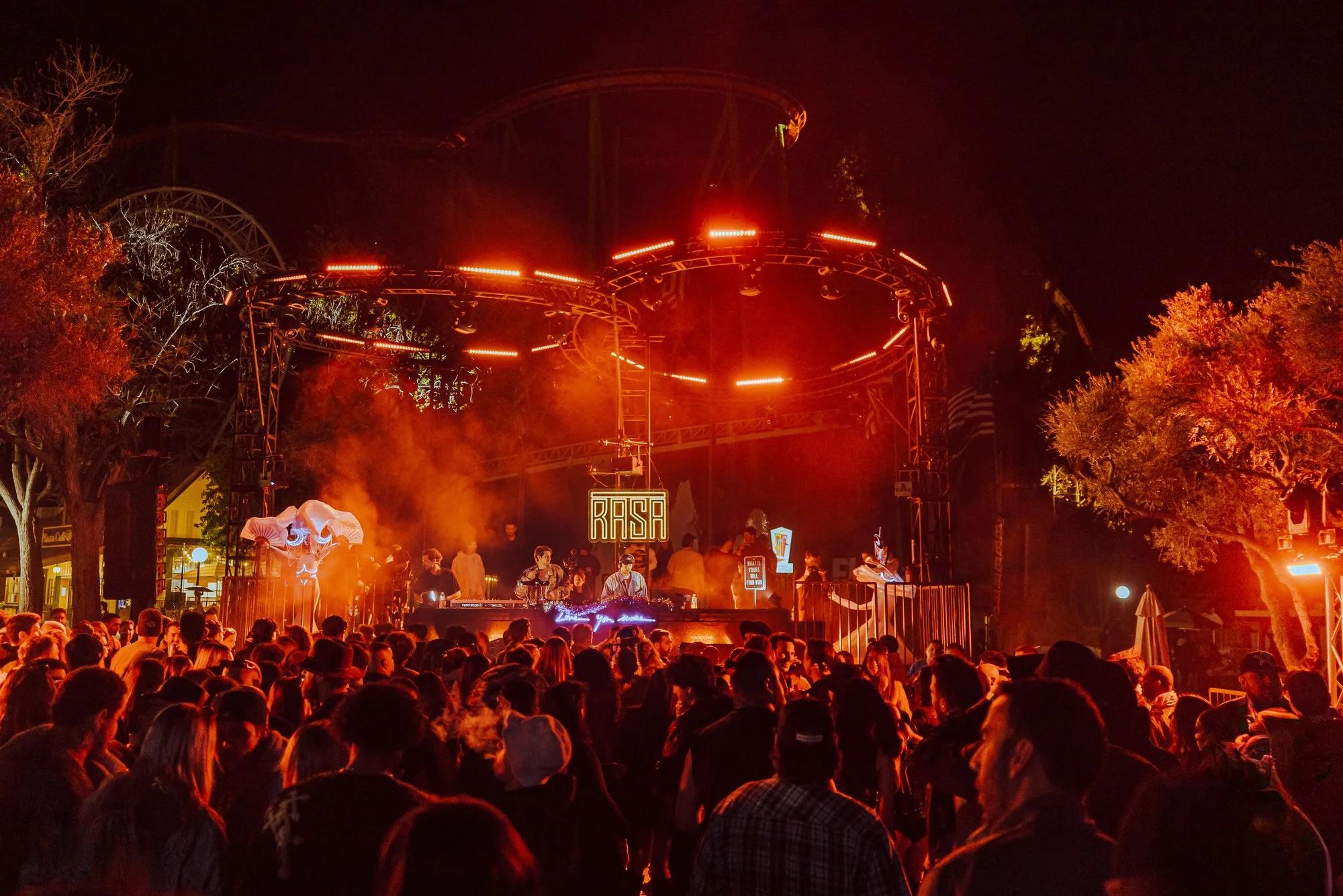 Red-lit circular truss ring over a DJ booth with RASA sign, crowd in warm haze below